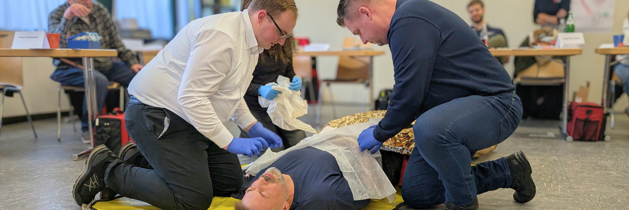 Szene aus einem Erste Hilfe Kurs: Ein Mann liegt am Boden. Zwei Männer knien neben ihm und leisten Erste Hilfe. Im Hintergrund zu sehen ist eine weitere Person, die Erste Hilfe leistet sowie mehrere zusehende Kursteilnehmende.