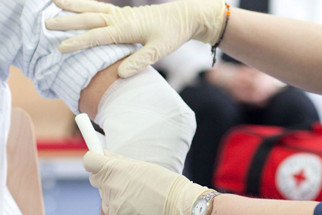 Verband wird am Arm angelegt. Im Hintergrund das Rote Kreuz Logo