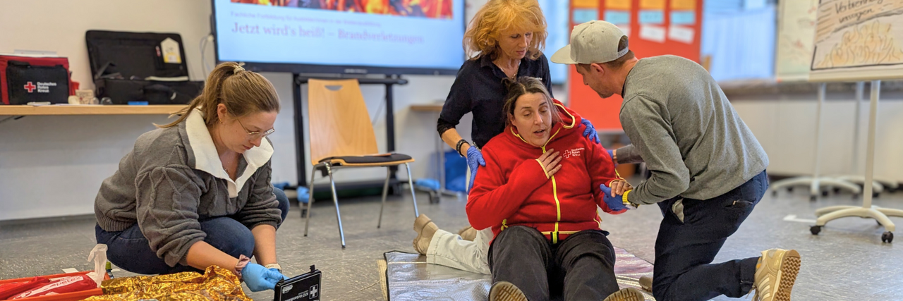 Szene aus einem Erste Hilfe Kurs: Eine Frau sitzt auf dem Boden, und bekommt scheinbar keine Luft. Eine Frau und ein Mann stützten sie. Eine weitere Frau holt etwas aus einem ERste Hilfe Koffer.