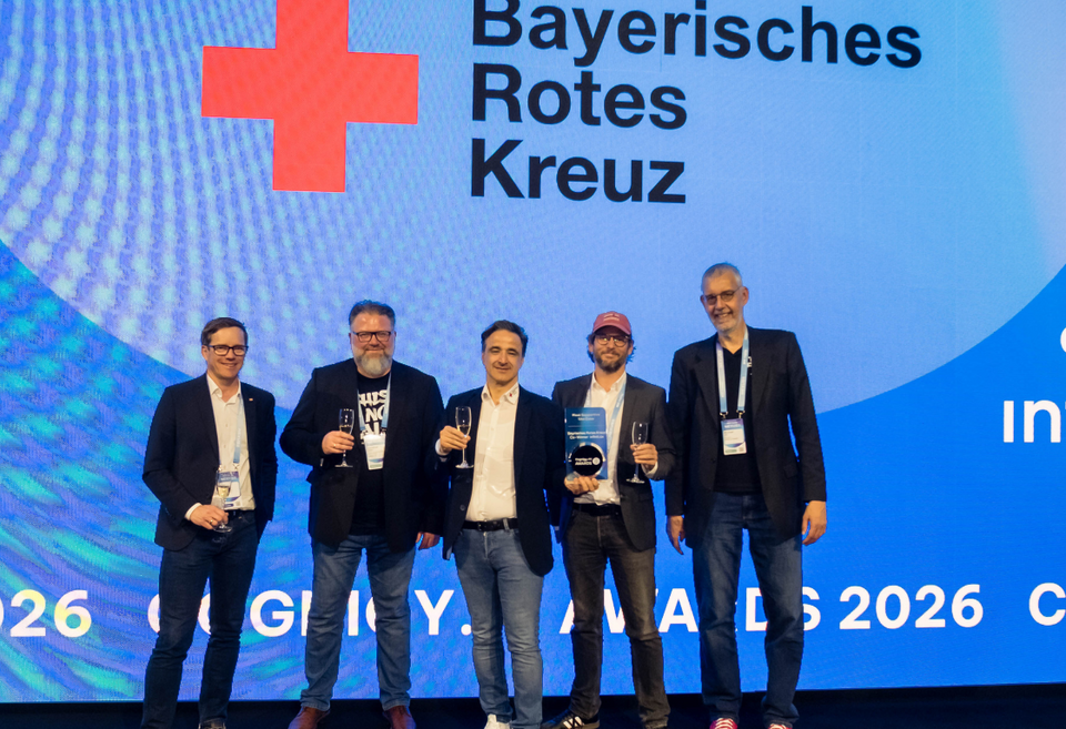 Foto von der Preisverleihung. Auf der Bühne stehen fünf Männer mit Sektgläsern in den Händen, in der Mitte Orhan Türkan, der Mann neben ihm hält den Preis auf Glas in den Händen. Im Hintergrund ist das BRK-Logo zu sehen.