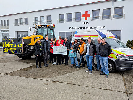 Gruppenfoto der Spendenübergabe: Christian Kammerer (6.v.r.) und Vertreter des Großhändlers OMEGA SORG überreichten im BRK-Katastrophenschutzzentrum in Atzenhof den Spendenscheck an Landrat Bernd Obst, BRK-Kreisgeschäftsführer Thomas Leipold (3. v.l.), stv. BRK-Kreisgeschäftsführerin Katrin Rohm (2.v.l.) sowie Herzenswunschinitiatorin Waltraud Heiter (5.v.l) und ihr ehrenamtliches Team., die gemeinsam vor dem Herzenswunschmobil und einem geschmückten Traktor stehen. Im Hintergrund ist das Gebäude des BRK-Katastrophenschutzzentrums in Atzenhof zu sehen.