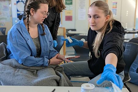 Foto von der Station Erste Hilfe Praxis beim Kreiswettbewerb: Ein Unfall im Chemie-Labor war das fiktive Szenario: Eine junge Frau (rechts) verbindet eine – täuschend echt geschminke – Wunde an der Hand einer anderen jungen Frau im Labor-Kittel.