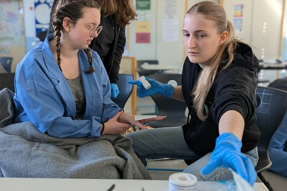 Foto von der Station Erste Hilfe Praxis beim Kreiswettbewerb: Ein Unfall im Chemie-Labor war das fiktive Szenario: Eine junge Frau (rechts) verbindet eine – täuschend echt geschminke – Wunde an der Hand einer anderen jungen Frau im Labor-Kittel.