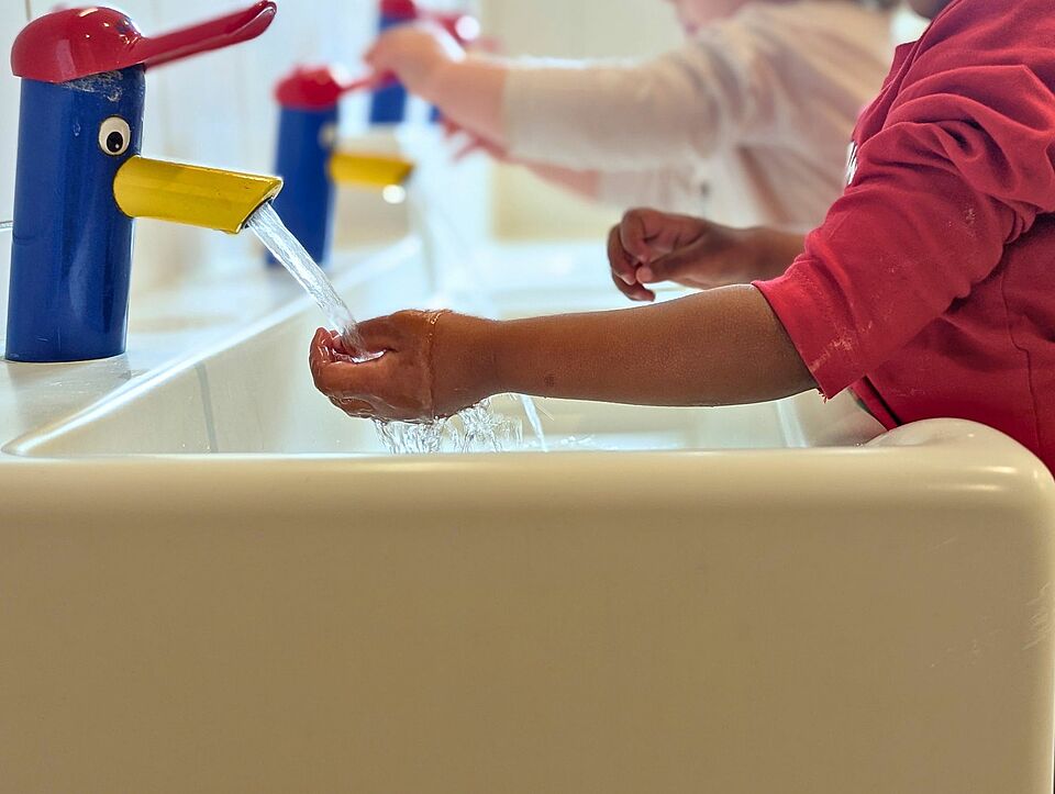 Zwei Kinder waschen sich die Hände, die Wasserhähne sind wie Enten gestaltet, die eine rote Kappe tragen.