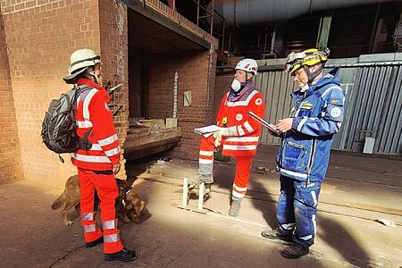 In der alten Ziegelei in Langenzenn stehen drei Personen und ein Hund vor einem alten Brennofen: Eine Frau in Rotkreuzeinsatzkleidung und schwarzem Rucksack mit einem Hund an der Leine, ihr gegebenüber die beiden Prüfer mit Klemmbrettern in der Hand. Einer von ihnen trägt Einsatzkleidung des BRK, der andere des THW.