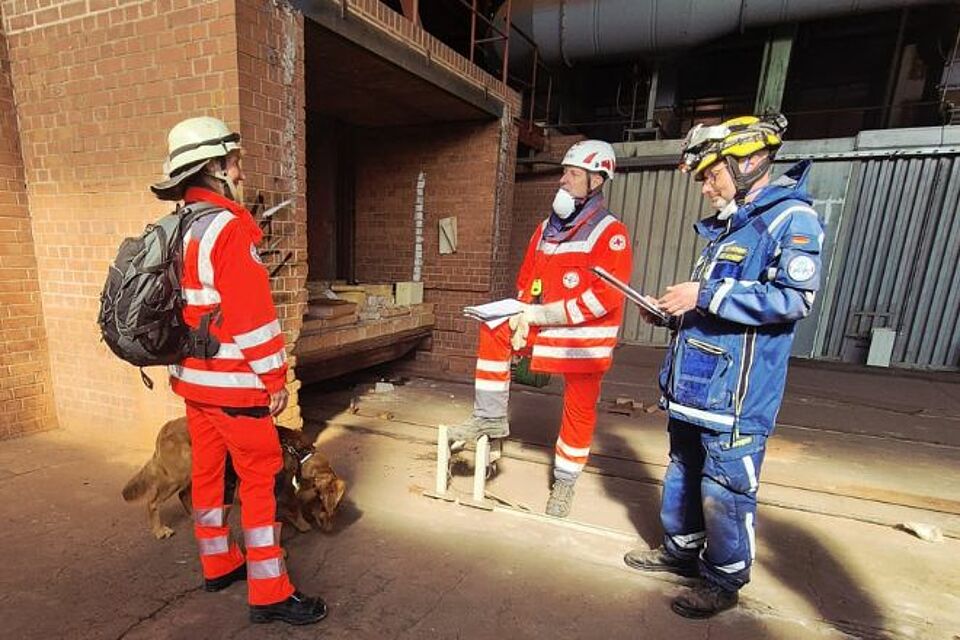 In der alten Ziegelei in Langenzenn stehen drei Personen und ein Hund vor einem alten Brennofen: Eine Frau in Rotkreuzeinsatzkleidung und schwarzem Rucksack mit einem Hund an der Leine, ihr gegebenüber die beiden Prüfer mit Klemmbrettern in der Hand. Einer von ihnen trägt Einsatzkleidung des BRK, der andere des THW.