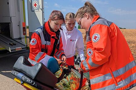 Drei junge Frauen in Rotkreuzeinsatzkleidung schnallen eine verunfallte Taucherin auf der Rettungstrage an.