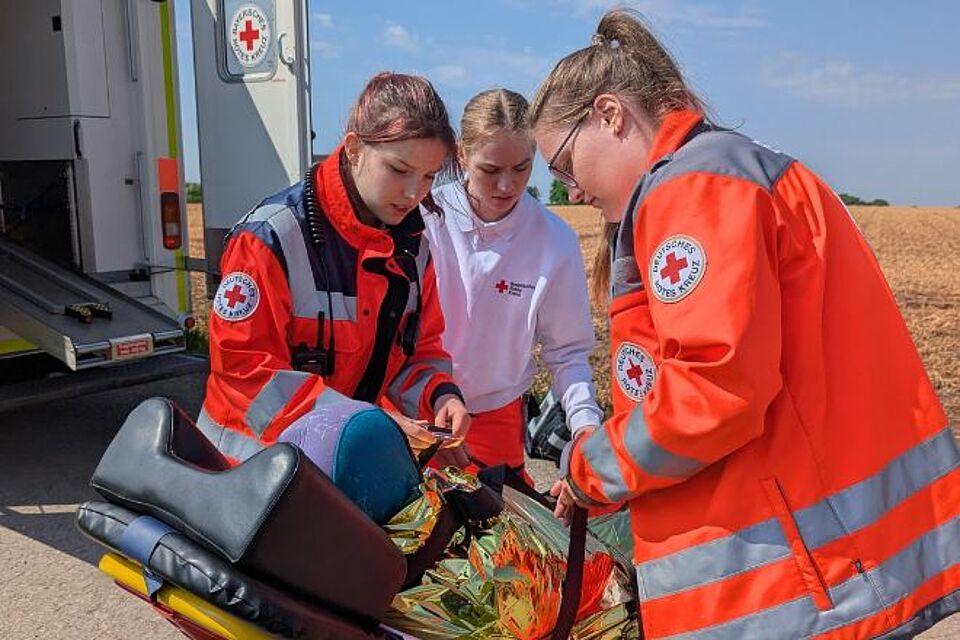 Drei junge Frauen in Rotkreuzeinsatzkleidung schnallen eine verunfallte Taucherin auf der Rettungstrage an.