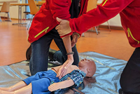 Frau führt Herzdruckmassage an Kinderpuppe vor. Mann korrigiert Armhaltung