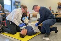Szene aus einem Erste Hilfe Kurs: Ein Mann liegt am Boden. Zwei Männer knien neben ihm und leisten Erste Hilfe. Im Hintergrund zu sehen ist eine weitere Person, die Erste Hilfe leistet sowie mehrere zusehende Kursteilnehmende.