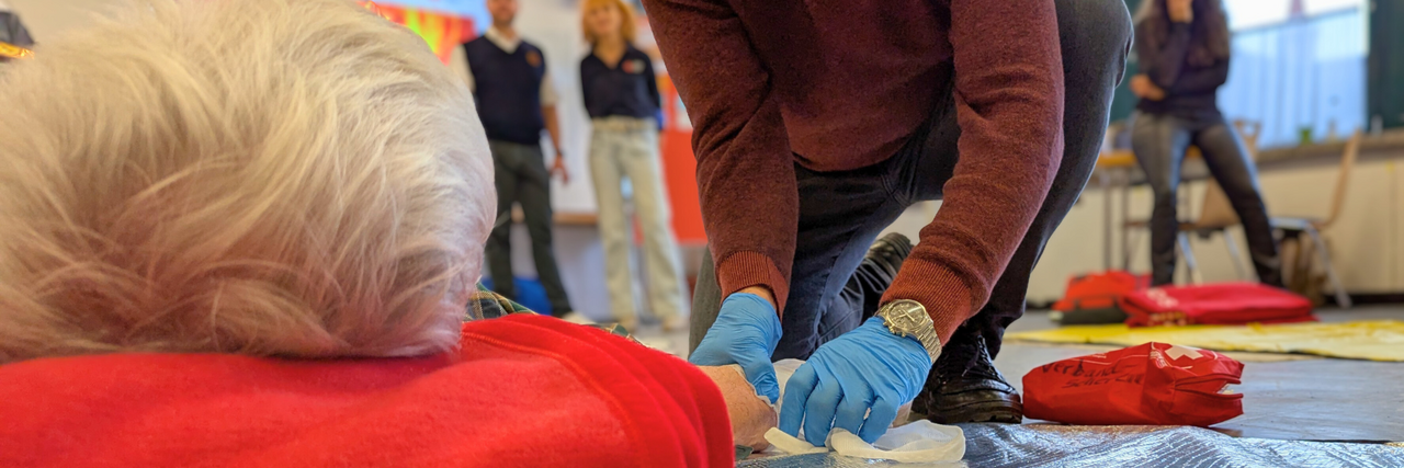 Szene aus einem Erste Hilfe Kurs: Nahaufnahme: Eine Hand legt einem Mann einen Verband am Arm an.