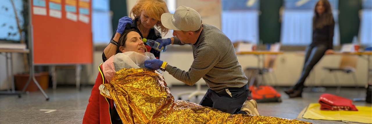 Szene aus einem Erste Hilfe Kurs: Eine Frau liegt am Boden und ist mit einer Goldfolie bedeckt, Eine Frau stützt sie, ein Mann gibt ihr Wasser aus einer Flasche.