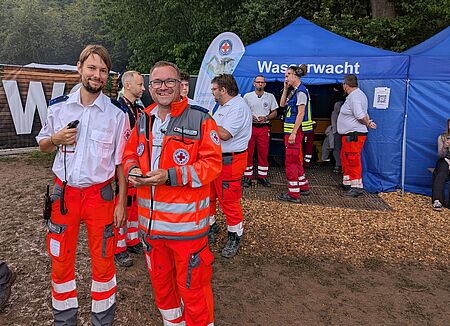 Zwei Sanitäter lächeln in die Kamera. Im Hintergrund zusehen ist das Festivalgelände des Open Beatz und weitere Sanitäter.