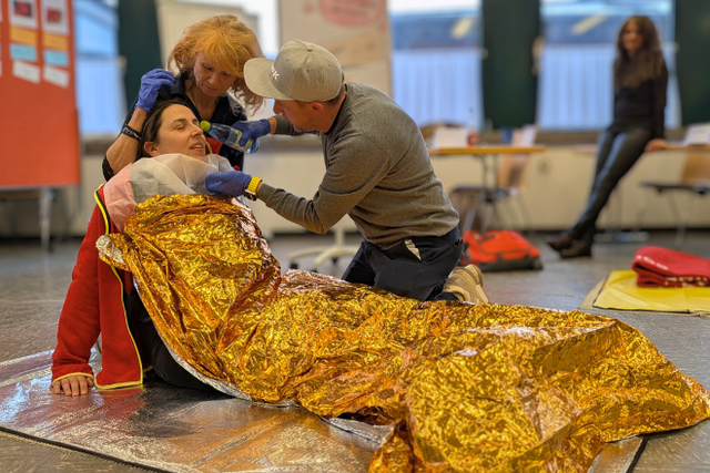 Szene aus einem Erste Hilfe Kurs: Eine Frau liegt am Boden und ist mit einer Rettungsdecke bedeckt, Eine Frau stützt sie, ein Mann gibt ihr Wasser aus einer Flasche.