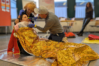 Szene aus einem Erste Hilfe Kurs: Eine Frau liegt am Boden und ist mit einer Rettungsdecke bedeckt, Eine Frau stützt sie, ein Mann gibt ihr Wasser aus einer Flasche.