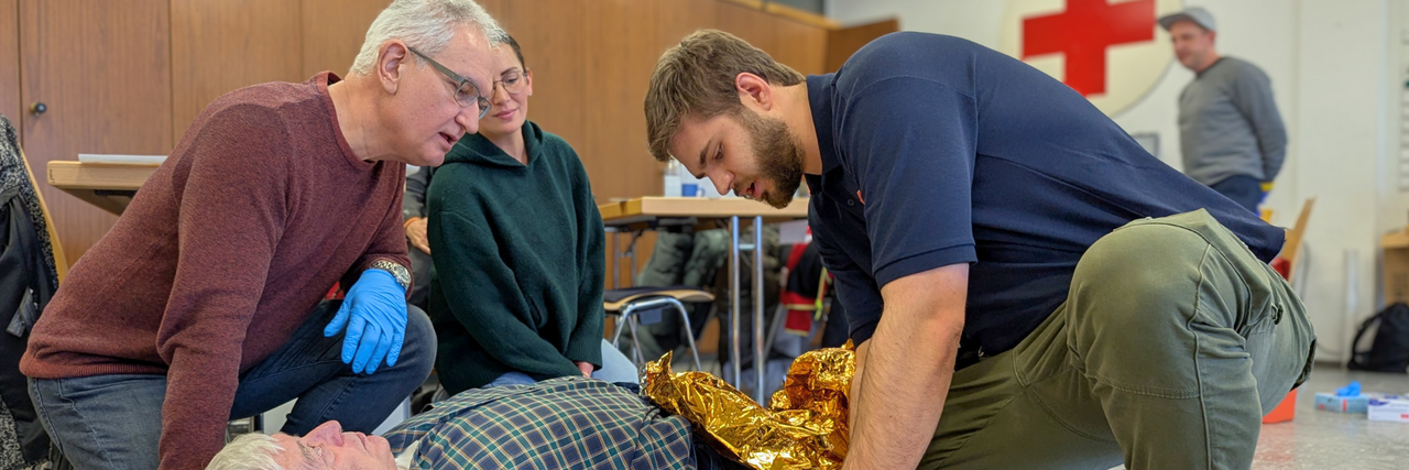 Szene eines Erste Hilfe Kurses: Ein Mann liegt am Boden, zwei Männer und eine Frau leiten Erste Hilfe.