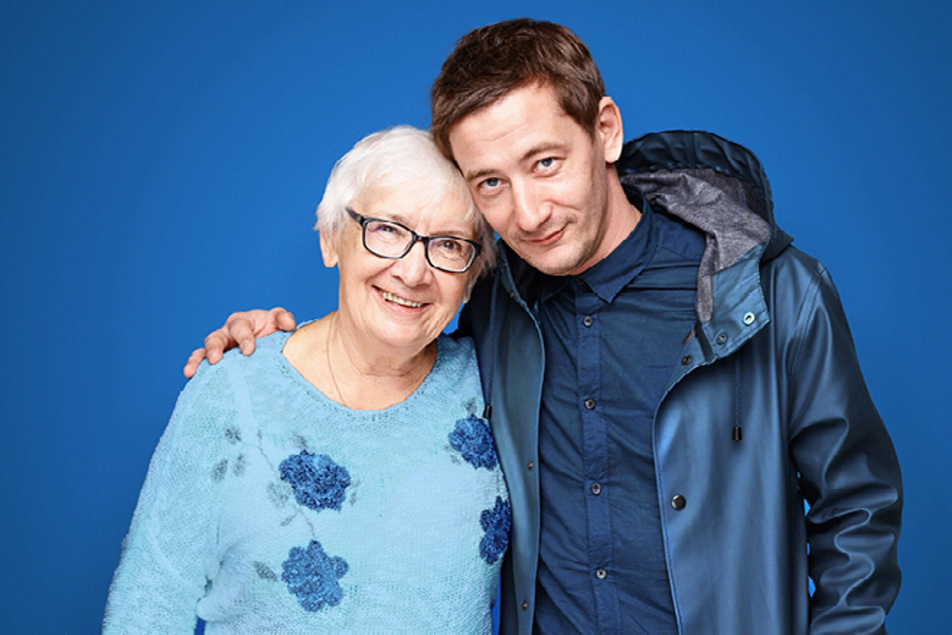 Erwachsener Mann legt Arm um seine Mutter. Hintergrund: Blau