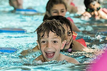 Foto: Ein Junge lernt mit viel Freude schwimmen. Im Hintergrund sind weitere Kinder im Schwimmbecken zu sehen.